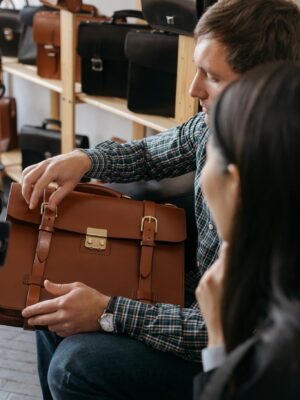 A man sits indoors examining a stylish leather briefcase in a store setting.