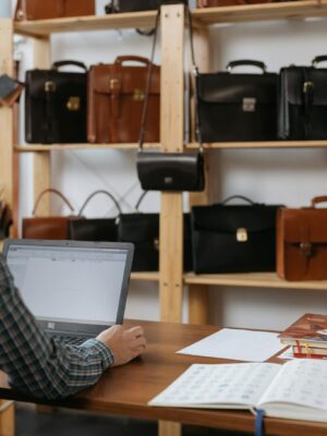 Businessman working on laptop in leather shop with briefcases on shelves.